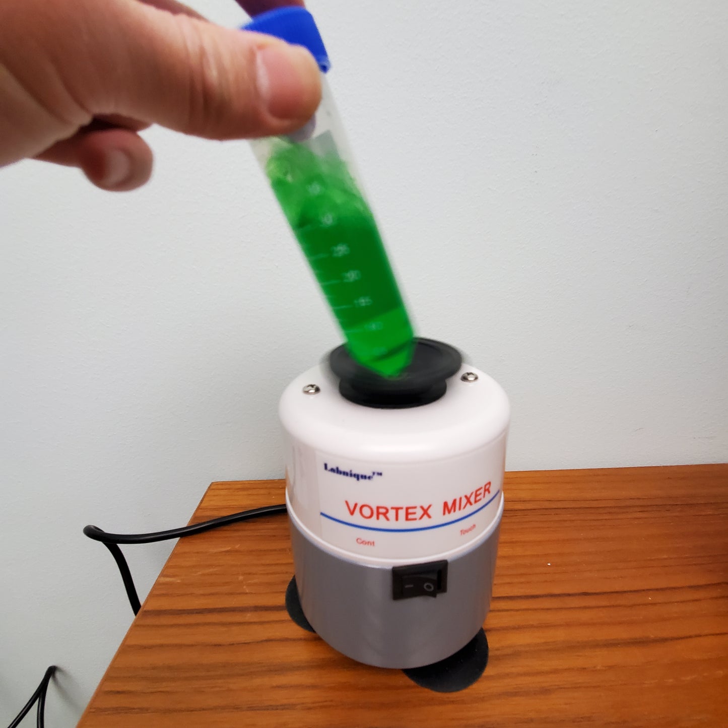 A Labnique vortex mixer with a white and gray body, black top, and black cord. A hand is placing a test tube with green liquid into the mixer. The mixer has a switch labeled "Cont" and "Touch." The mixer is on a wooden surface.