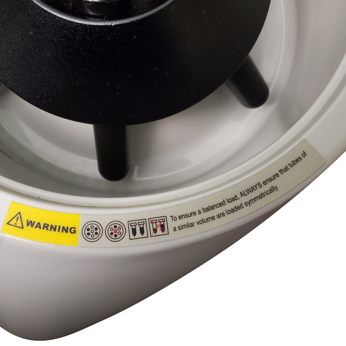 A close-up photograph of a white centrifuge rotor with a black rotor nut in the center. A yellow "WARNING" triangle is visible on the left, along with pictograms showing tube placement. Text to the right reads "To ensure a balanced load, ALWAYS ensure that tubes of a similar volume are loaded symmetrically." The image focuses on the safety warning and the rotor assembly.